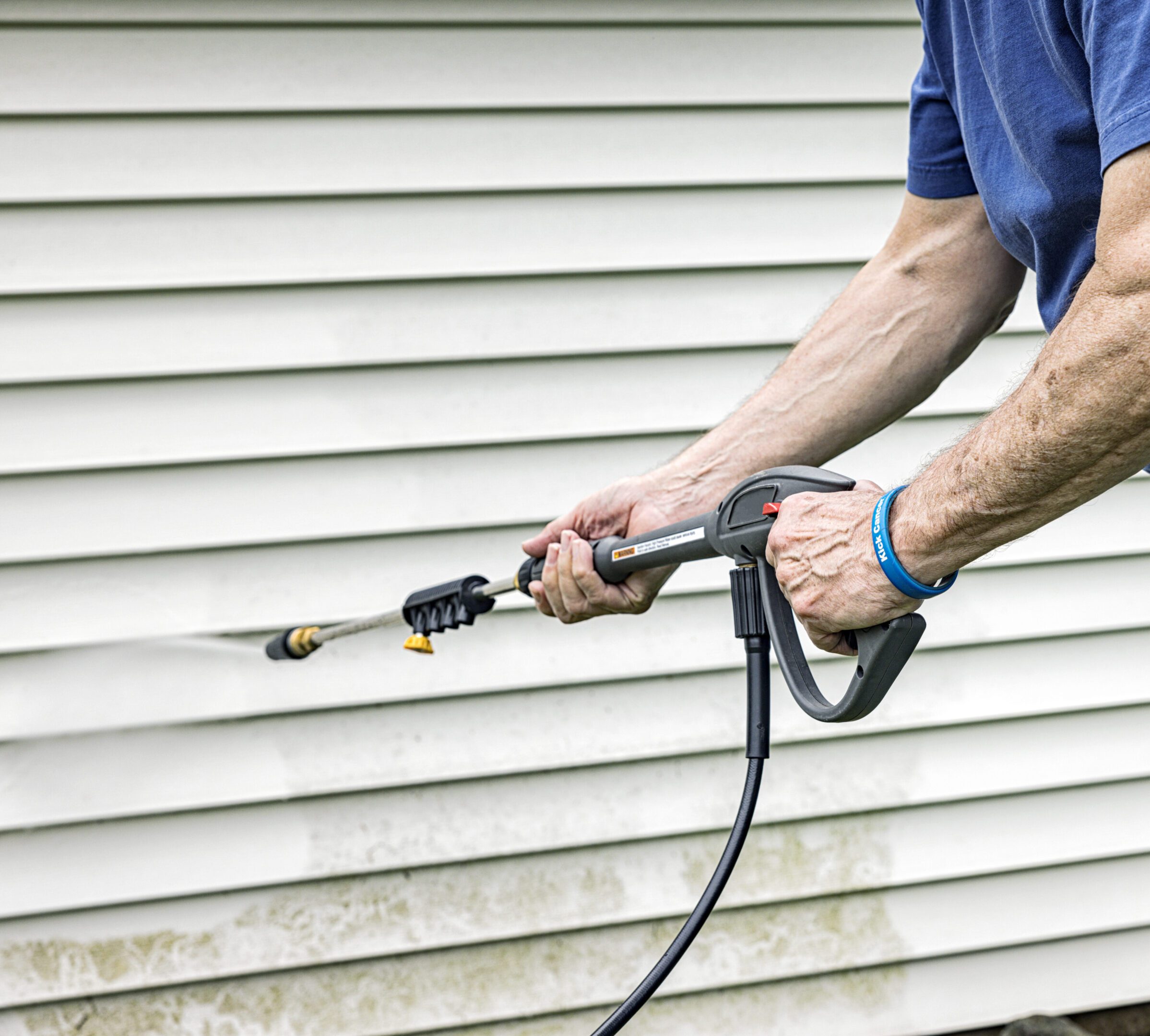 Real life, real person DIY senior adult man cancer survivor is cleaning a section of vinyl clapboard siding on the residential home garden shed with high pressure cleaning power wash equipment. He is wearing a self-designed (by the photographer/model/cancer survivor) wristband labeled "Kick Cancer" as he continues to stay as active as possible after surviving a few lousy years of cancer and chemotherapy "adventures".