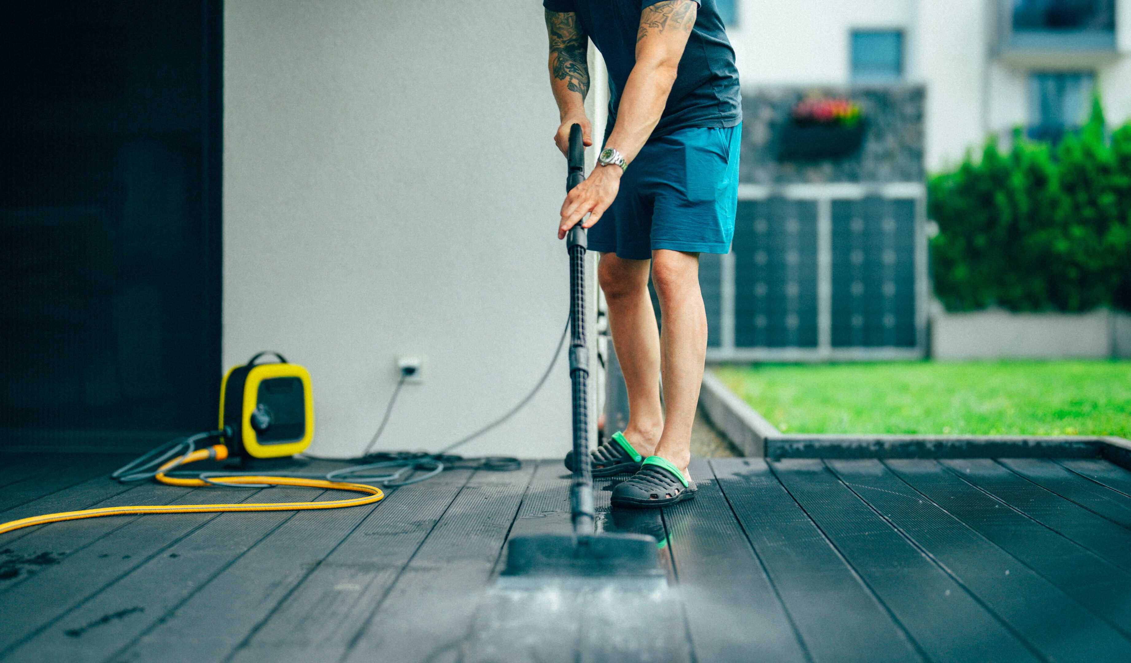 Man cleans an outdoor wooden terrace using a pressure washer in the backyard of a modern house. He is dressed in casual clothes. Solar panels are visible in the background beside a green lawn, highlighting sustainable living and home maintenance. The setting is contemporary and tidy, illustrating responsible outdoor cleaning and renewable energy features in a residential environment.